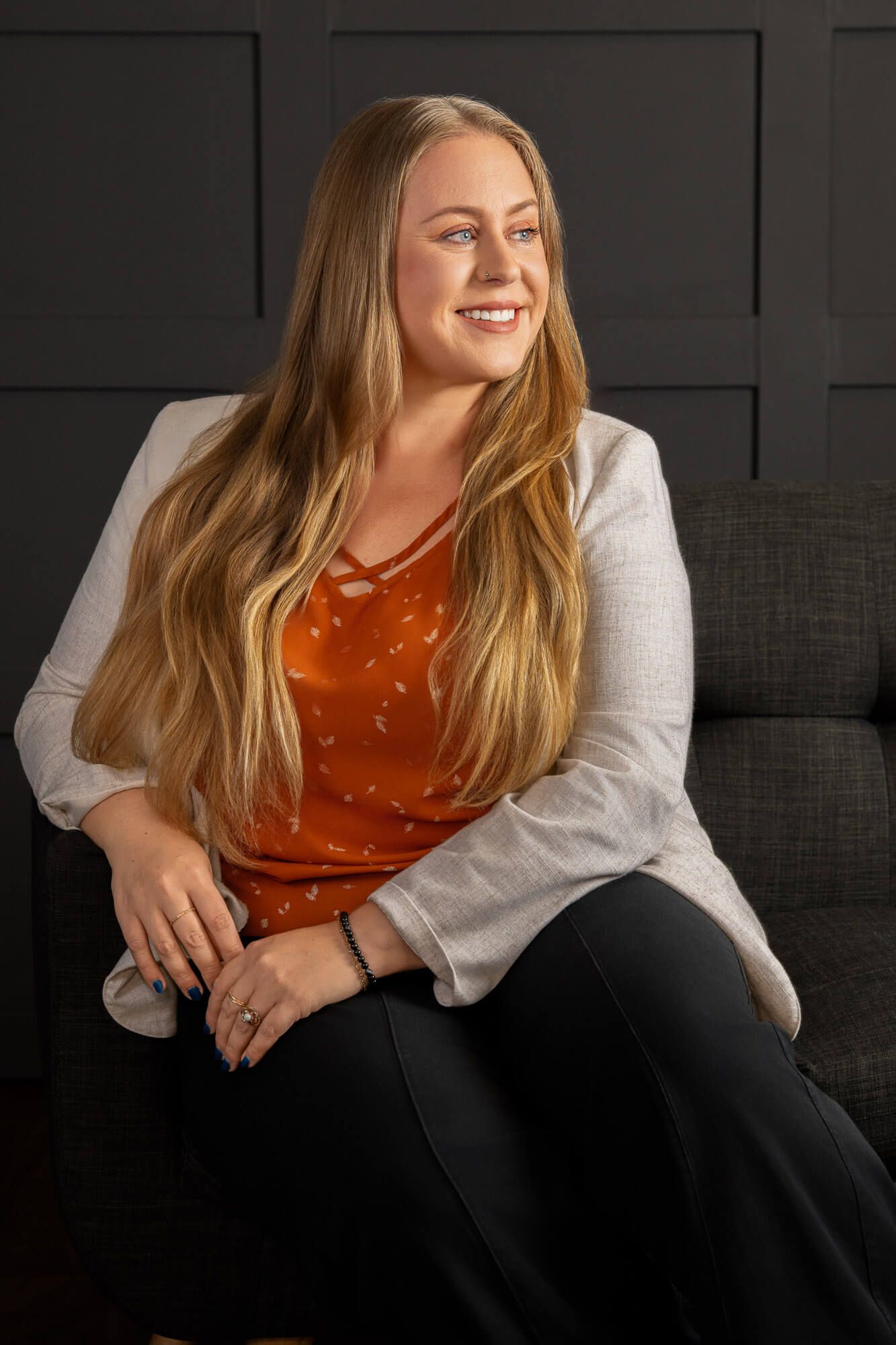 Casual portrait of someone in an orange top and white cardigan relaxing on a dark couch.