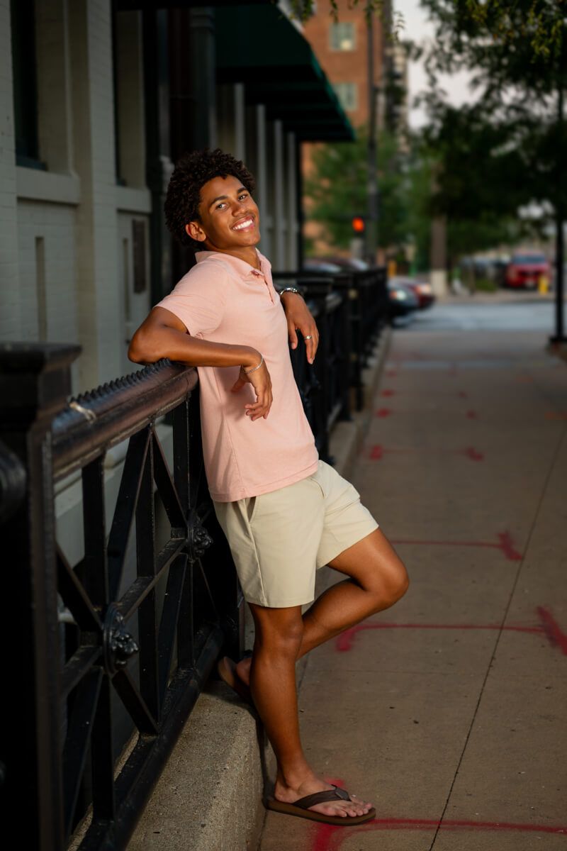 Smiling teen in pink polo and khaki shorts leaning on urban railing outdoors.