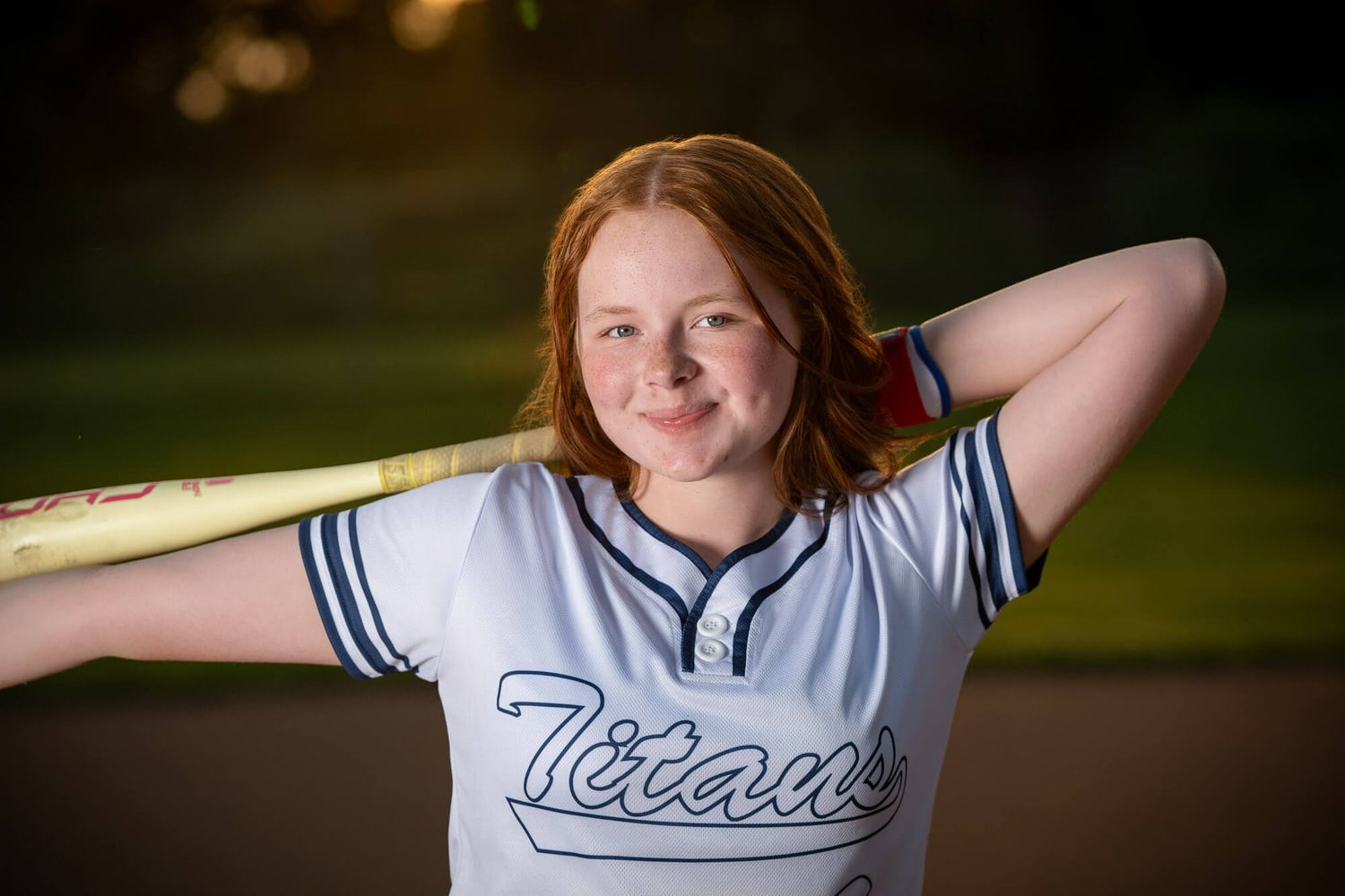 Young redhead girl in Titans softball jersey poses with bat behind shoulders on field.