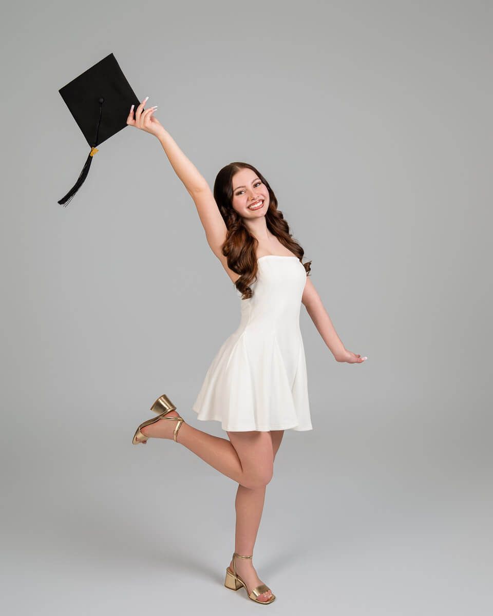 Smiling graduate in white dress holding black mortarboard cap up high, celebrating graduation against gray background.
