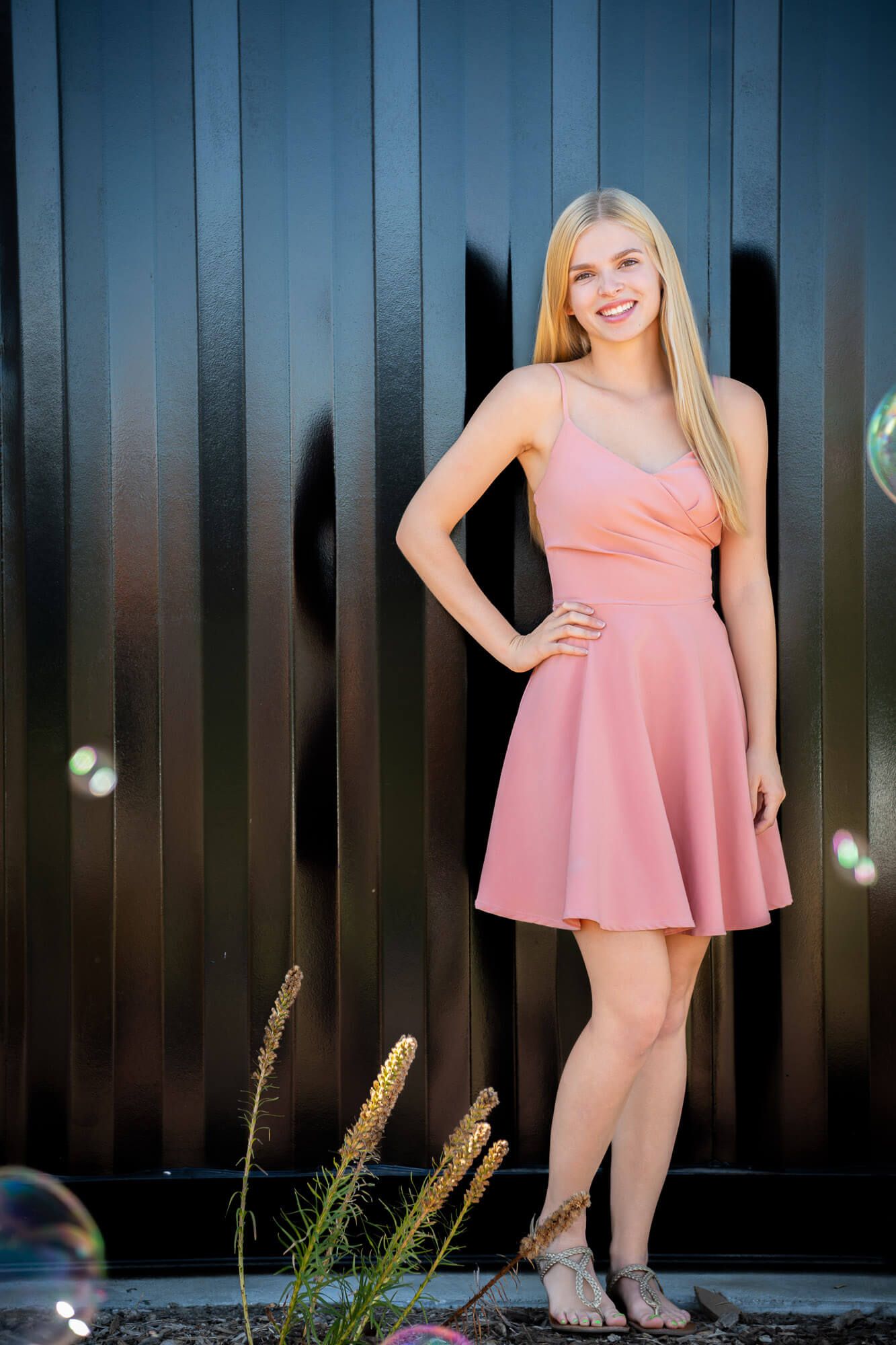 Young blonde woman in pink spaghetti strap dress posing confidently against dark metal wall.