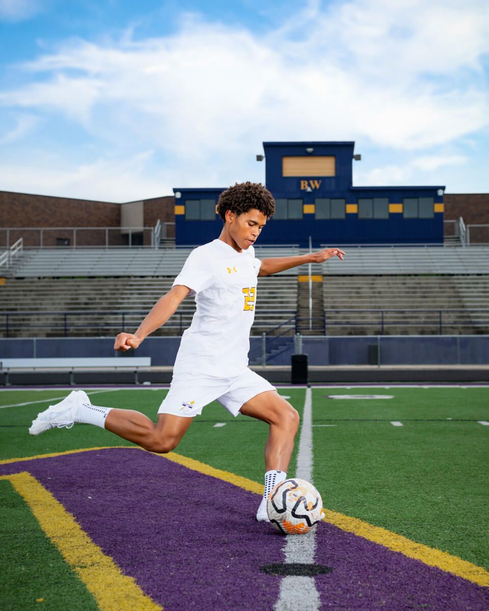 A young soccer player in white uniform kicks a ball on a purple and gold field with stadium bleachers behind him.