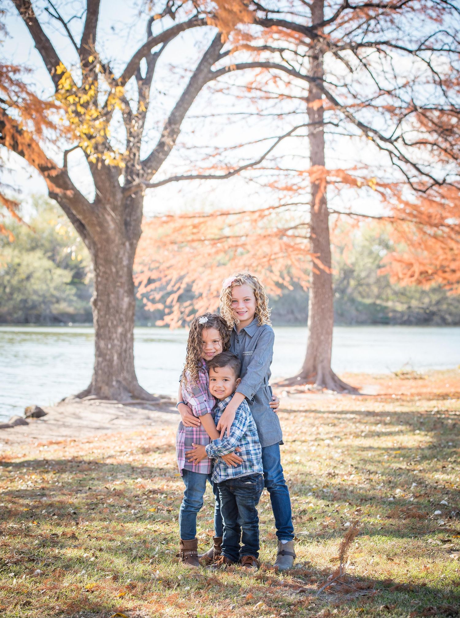 Children hugging together on an autumn day by the lake with fall foliage and golden sunlight during an Omaha family photography session.