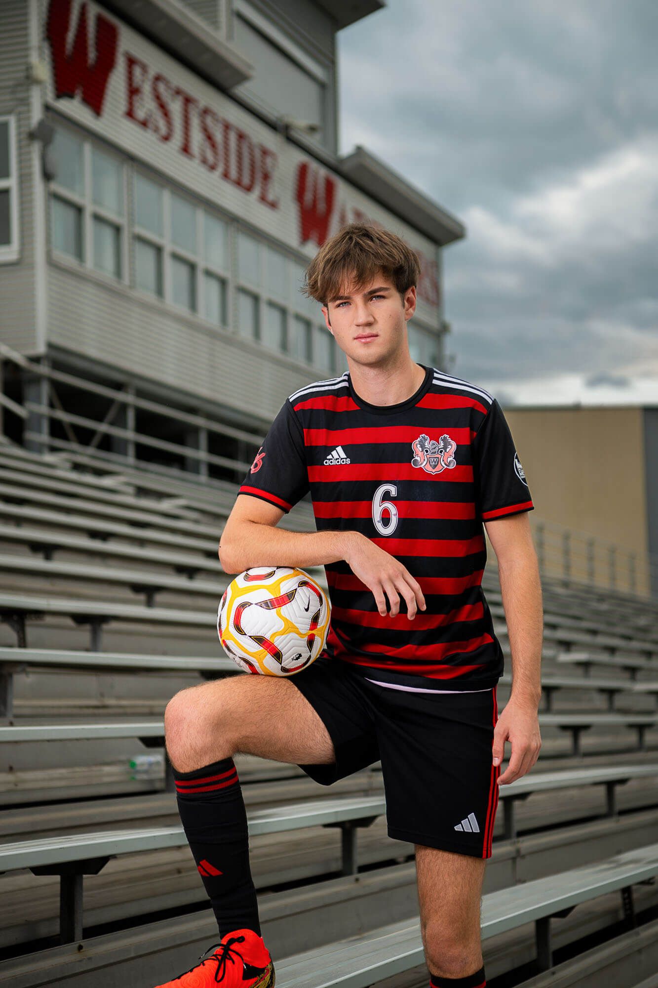 Soccer player in red and black striped jersey poses with ball on bleachers at Westside stadium.