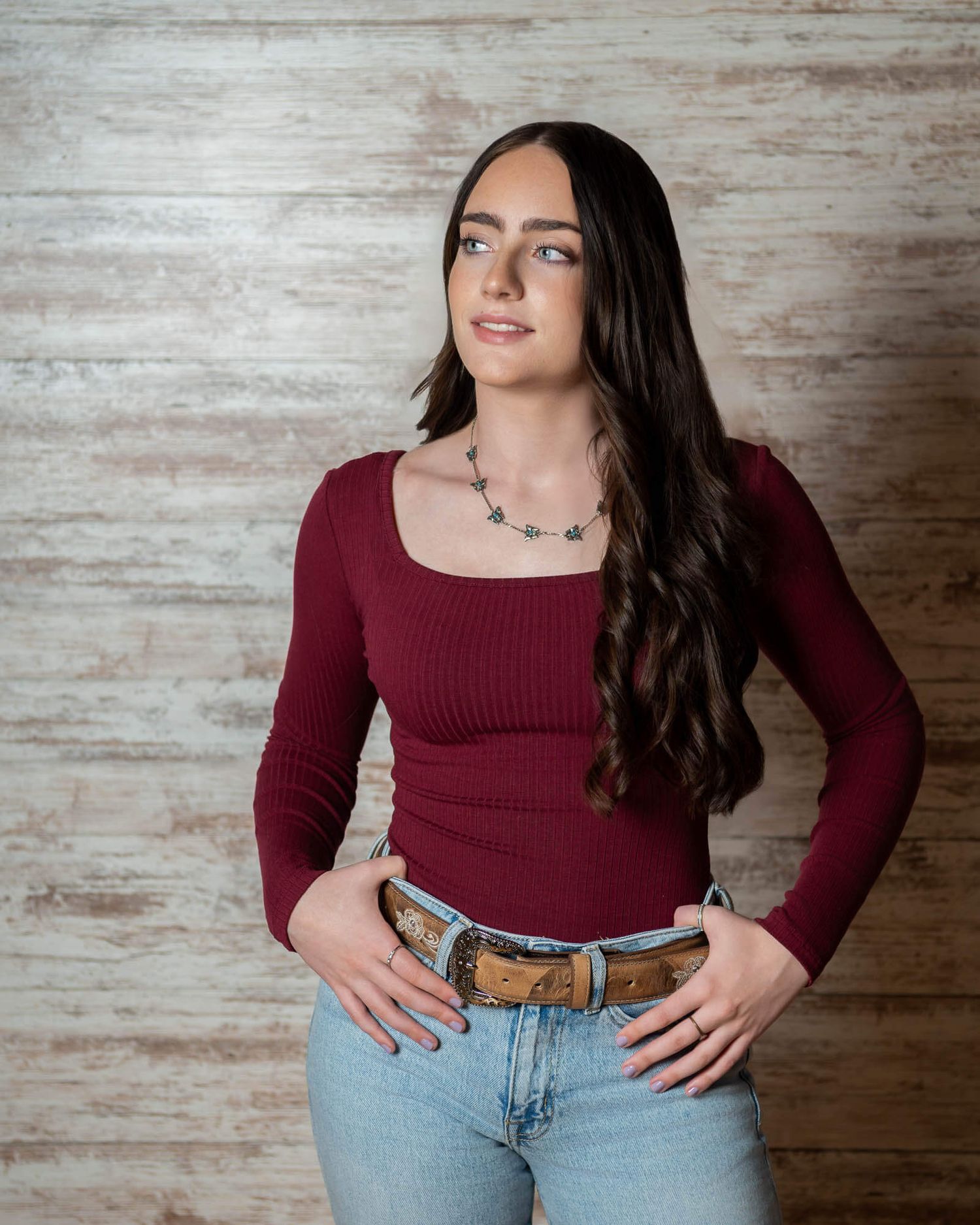 Woman in burgundy top and jeans poses casually against whitewashed wooden wall.