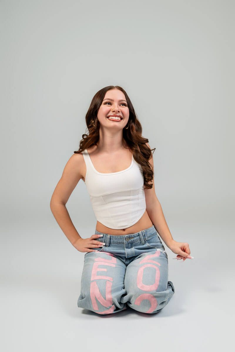 Smiling young woman in white crop top and graphic jeans sitting on gray background.