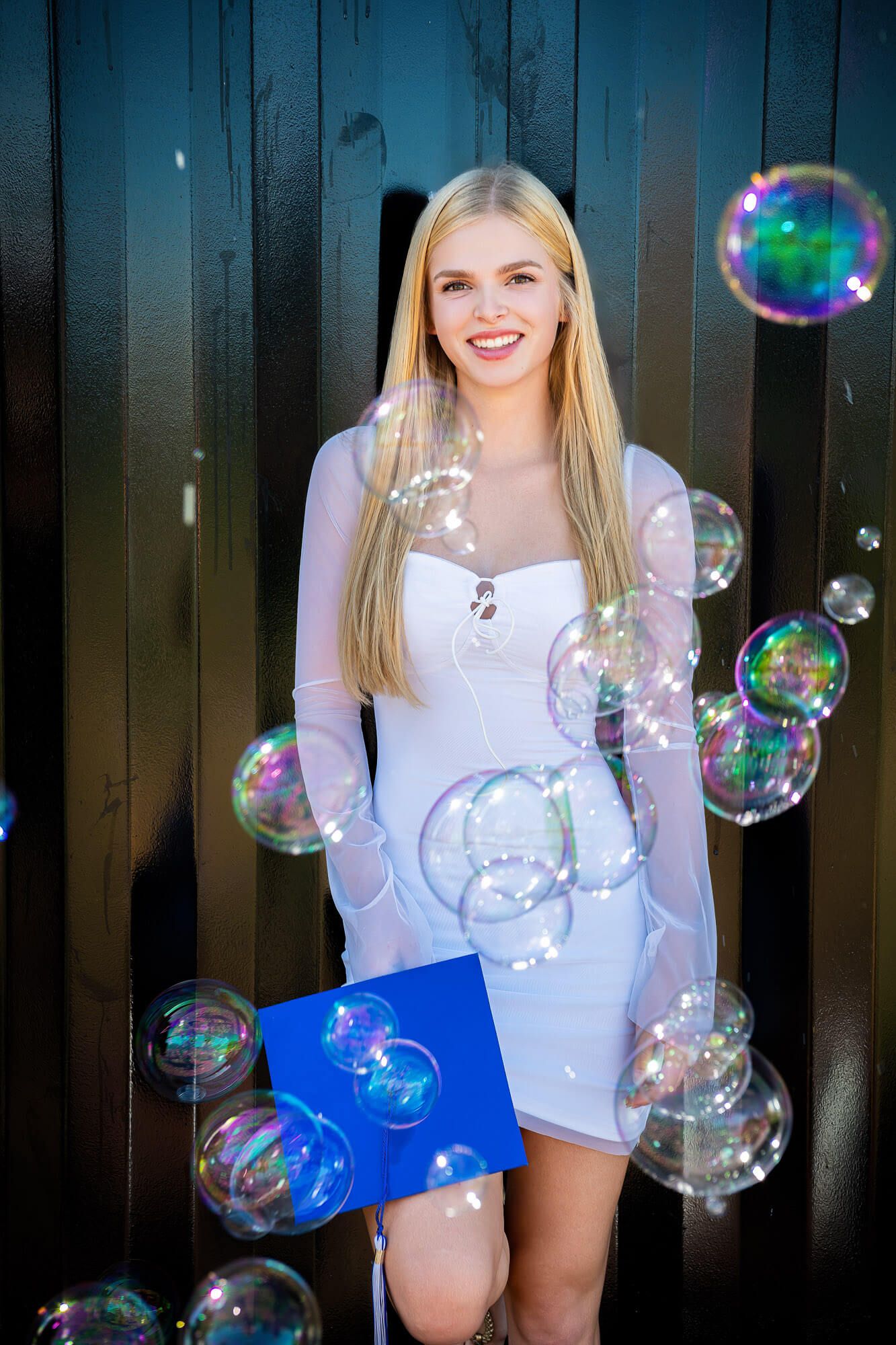 Cheerful blonde woman in white dress smiling while holding blue folder amid floating soap bubbles.