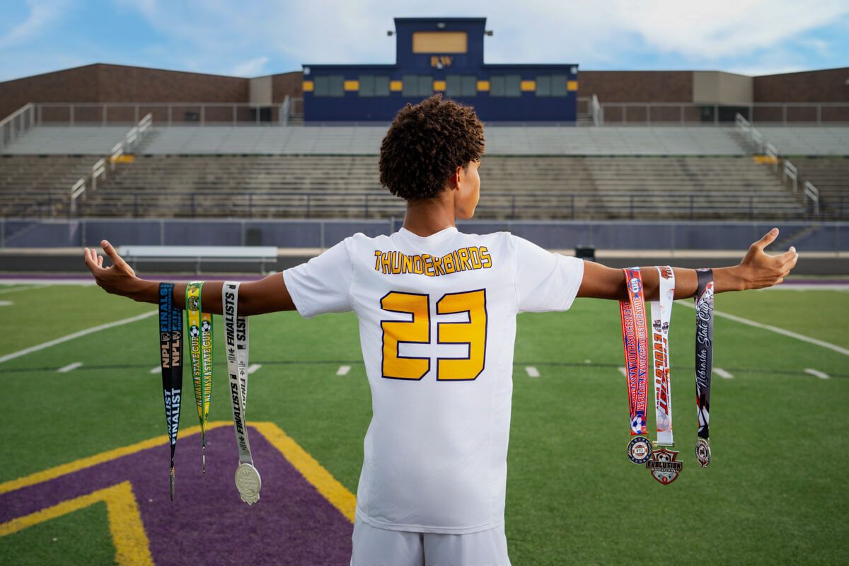 Teen athlete #23 displays medals and ribbons on a football field with arms outstretched, wearing a white jersey.