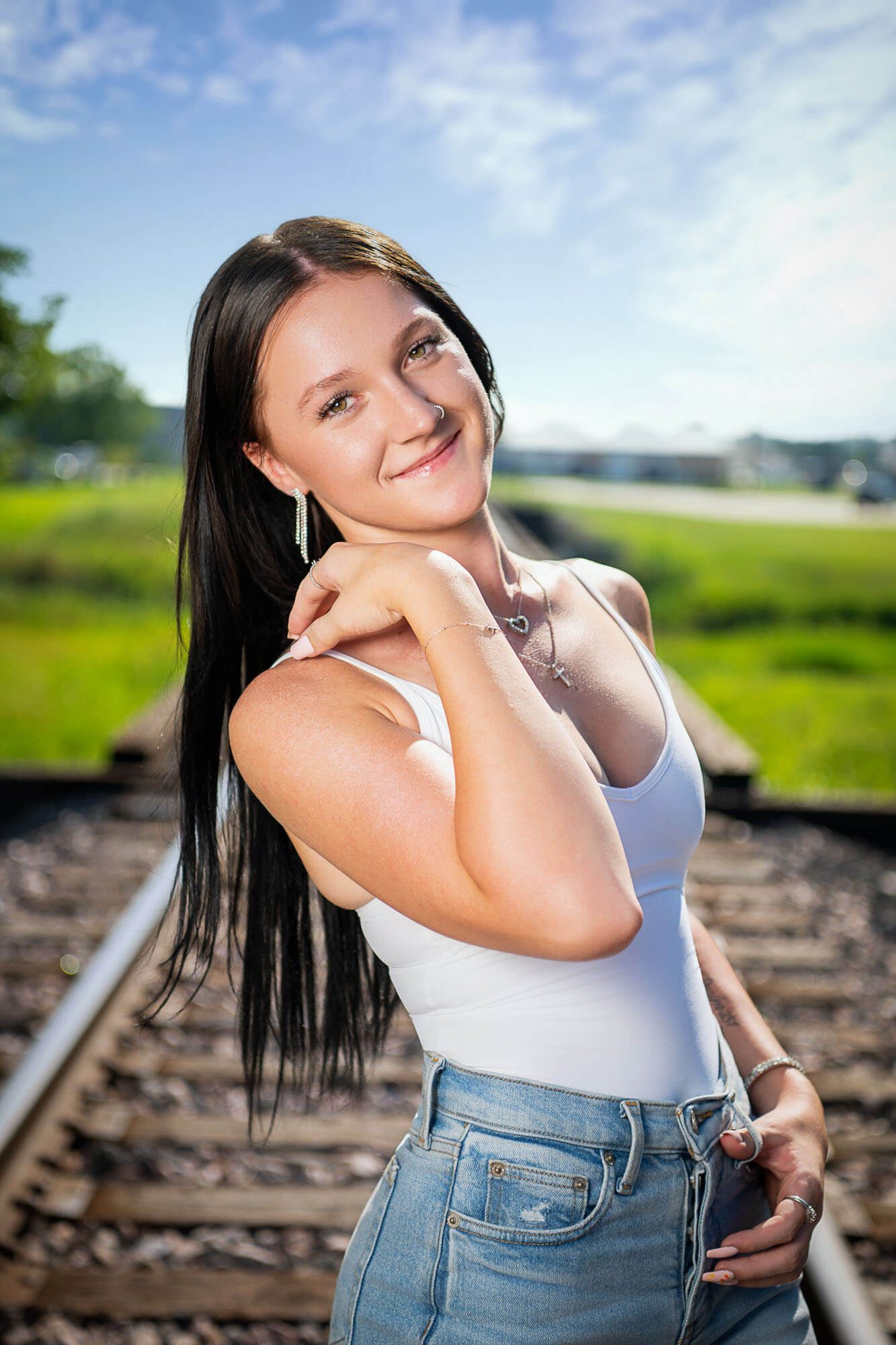 High school senior in Norfolk, NE, near Omaha, in a white tank top and jeans poses playfully on train tracks in a sunny outdoor setting.