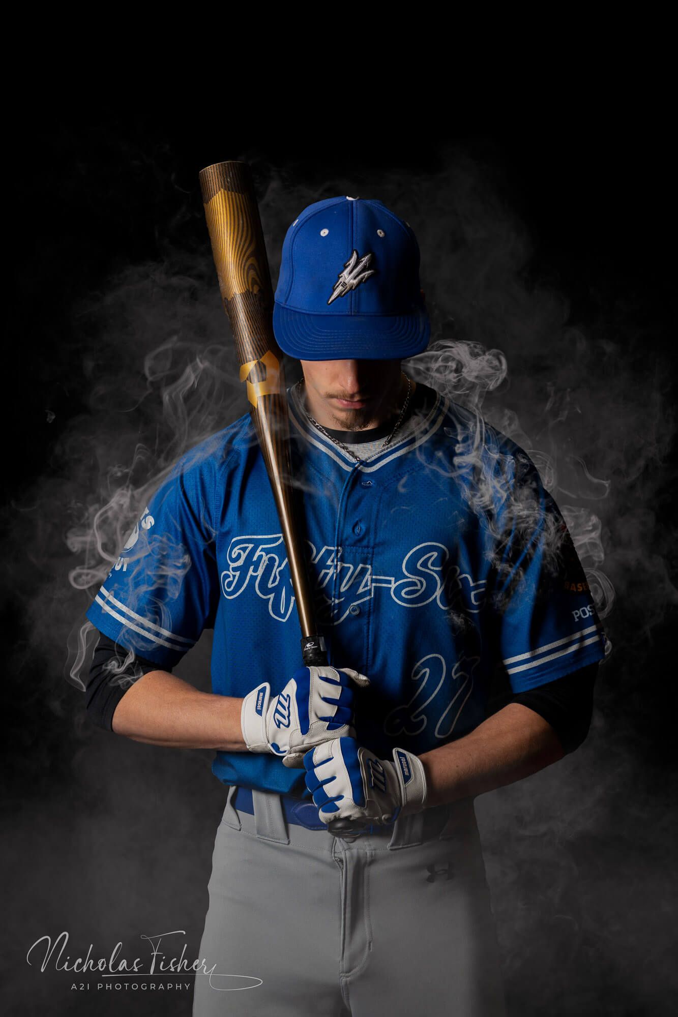 Baseball player in blue uniform posing with bat against dramatic smoky dark background.