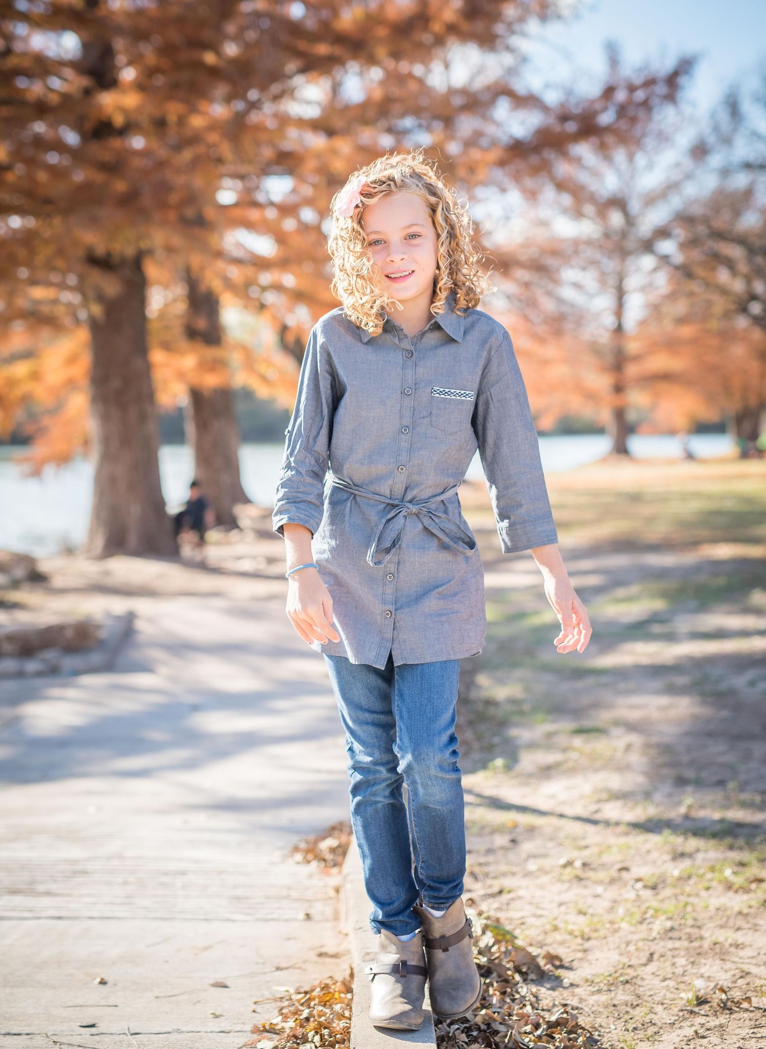 A girl stands on a sunlit path wearing a casual gray button-down shirt and jeans among autumn trees during family photography session.