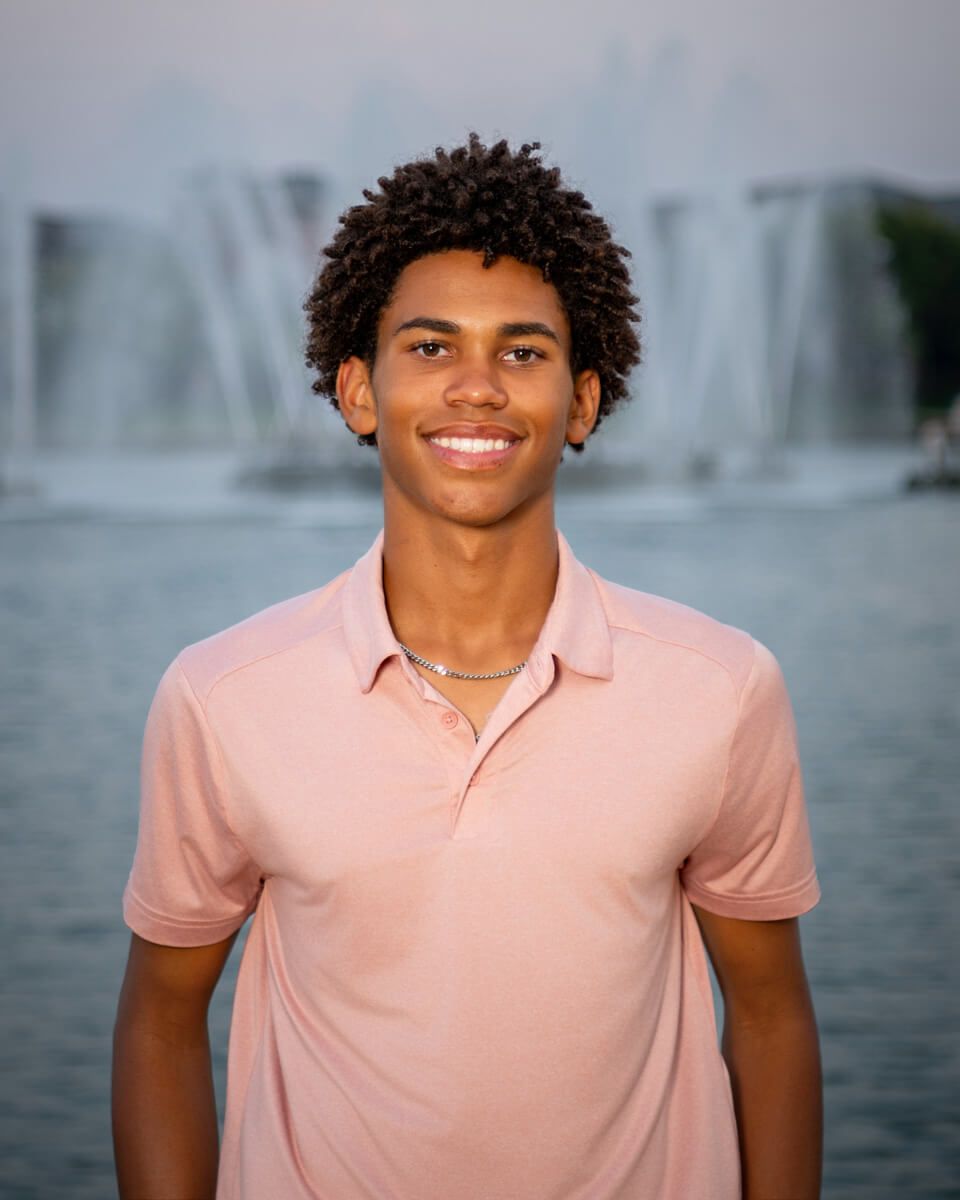 Smiling young man in pink polo shirt posing near fountain outdoors at dusk.