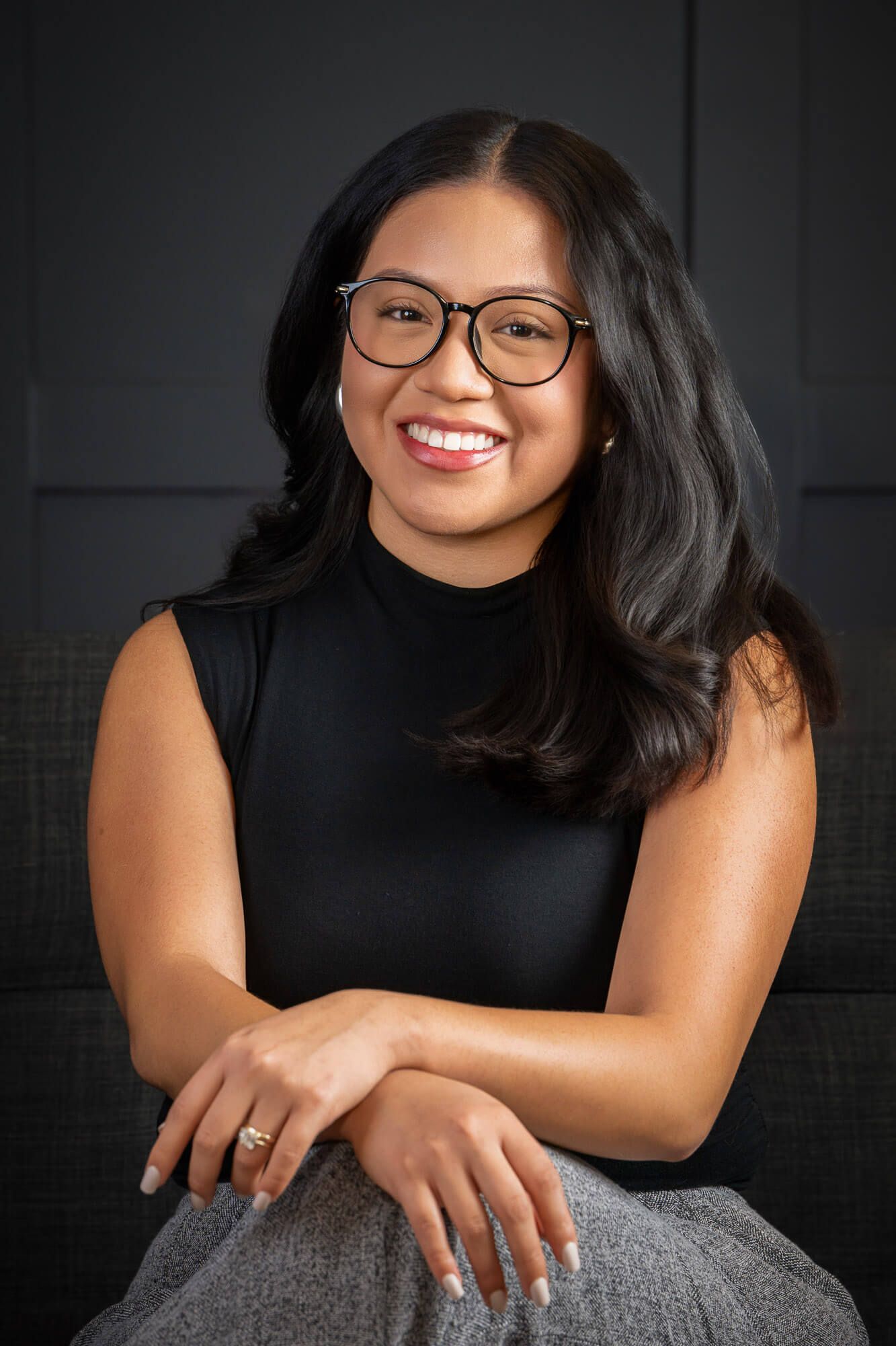 Professional portrait of someone in black sleeveless top and glasses smiling warmly against dark background.