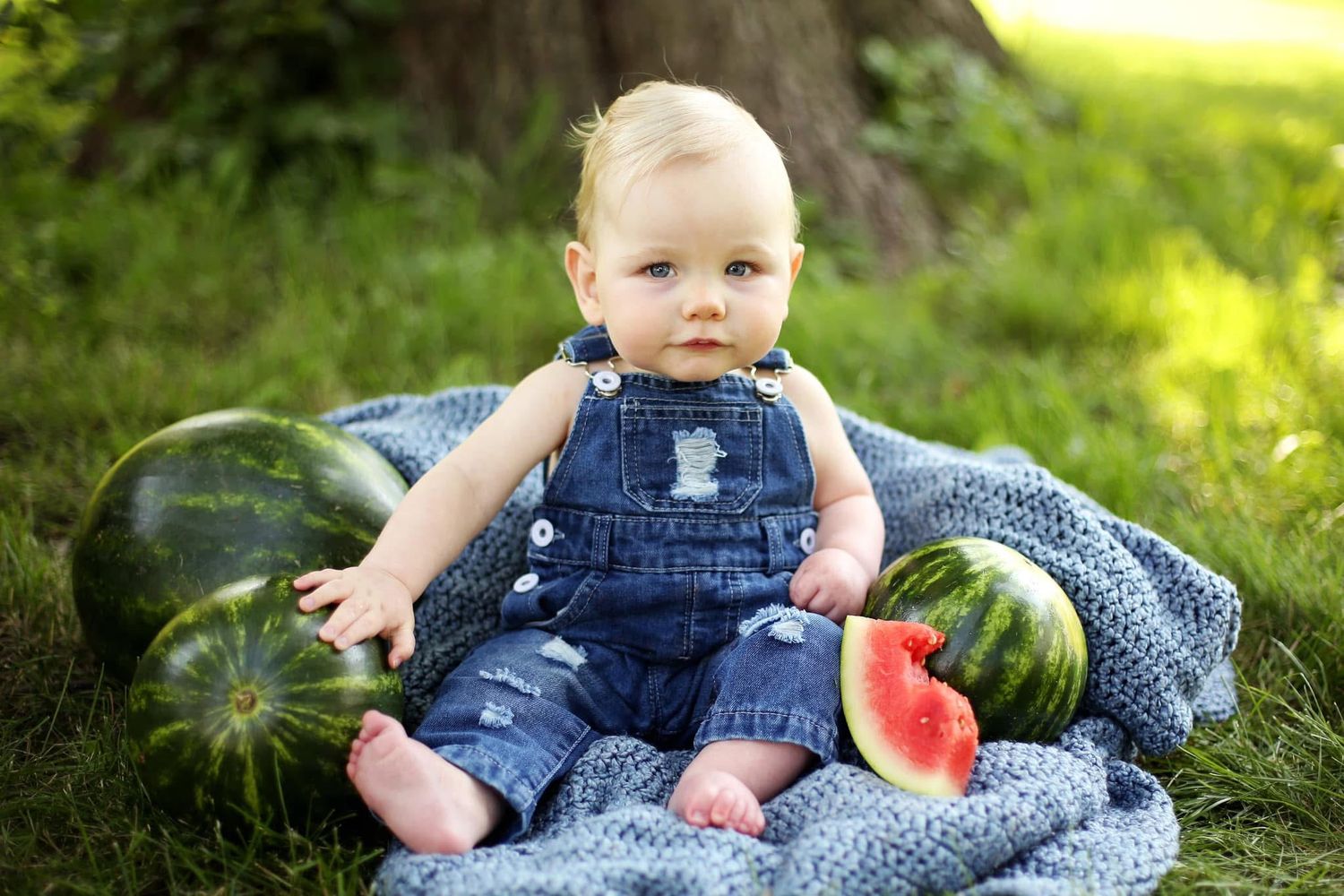 Young child in denim overalls sits on blue blanket outdoors with fresh watermelons on a summer day.