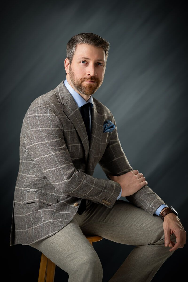 Bearded man in plaid blazer with blue pocket square sits confidently on stool against gray studio backdrop.