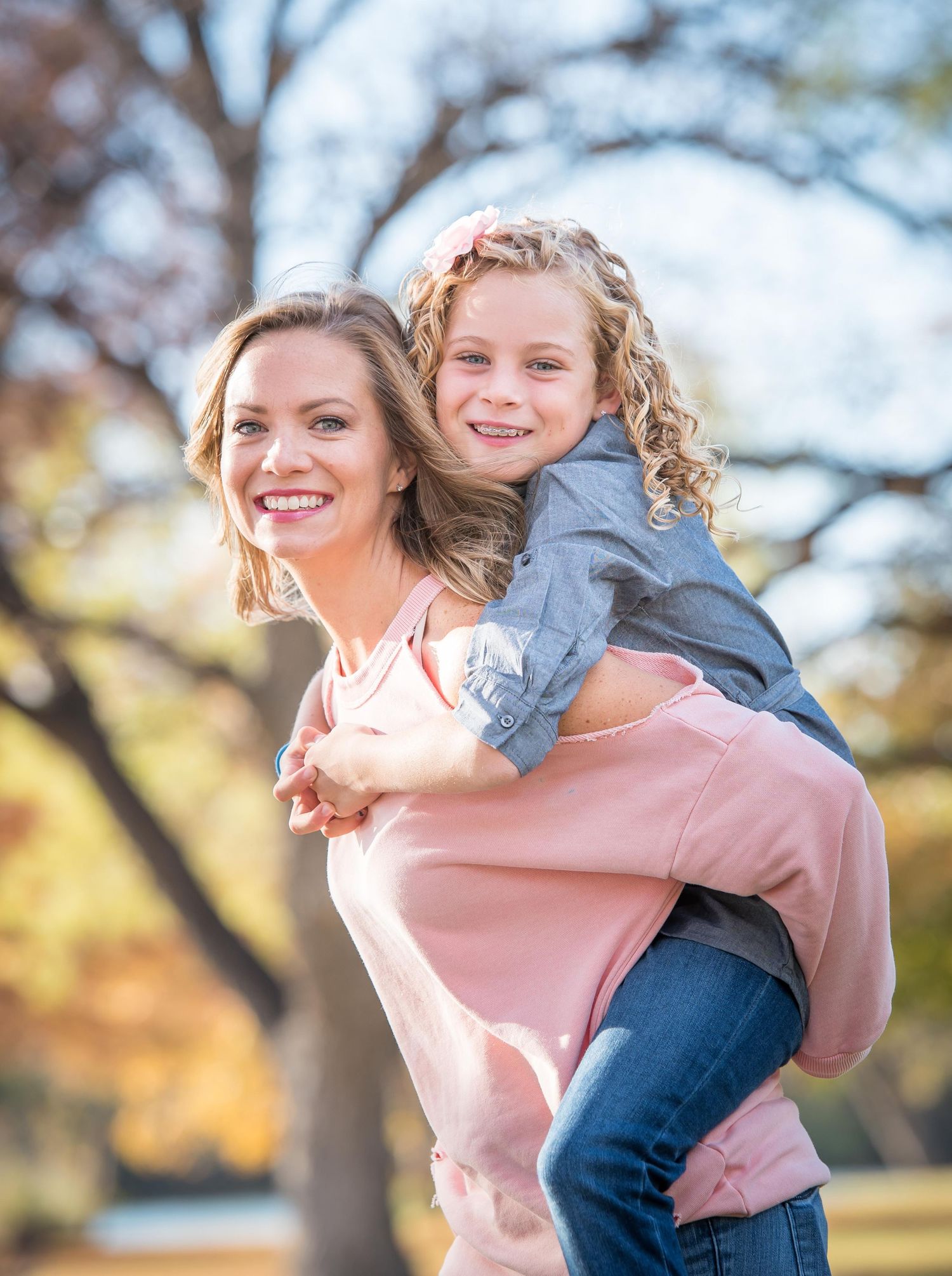 A joyful piggyback ride moment between mom and daughter outdoors on an autumn day with trees during Omaha family photography session.