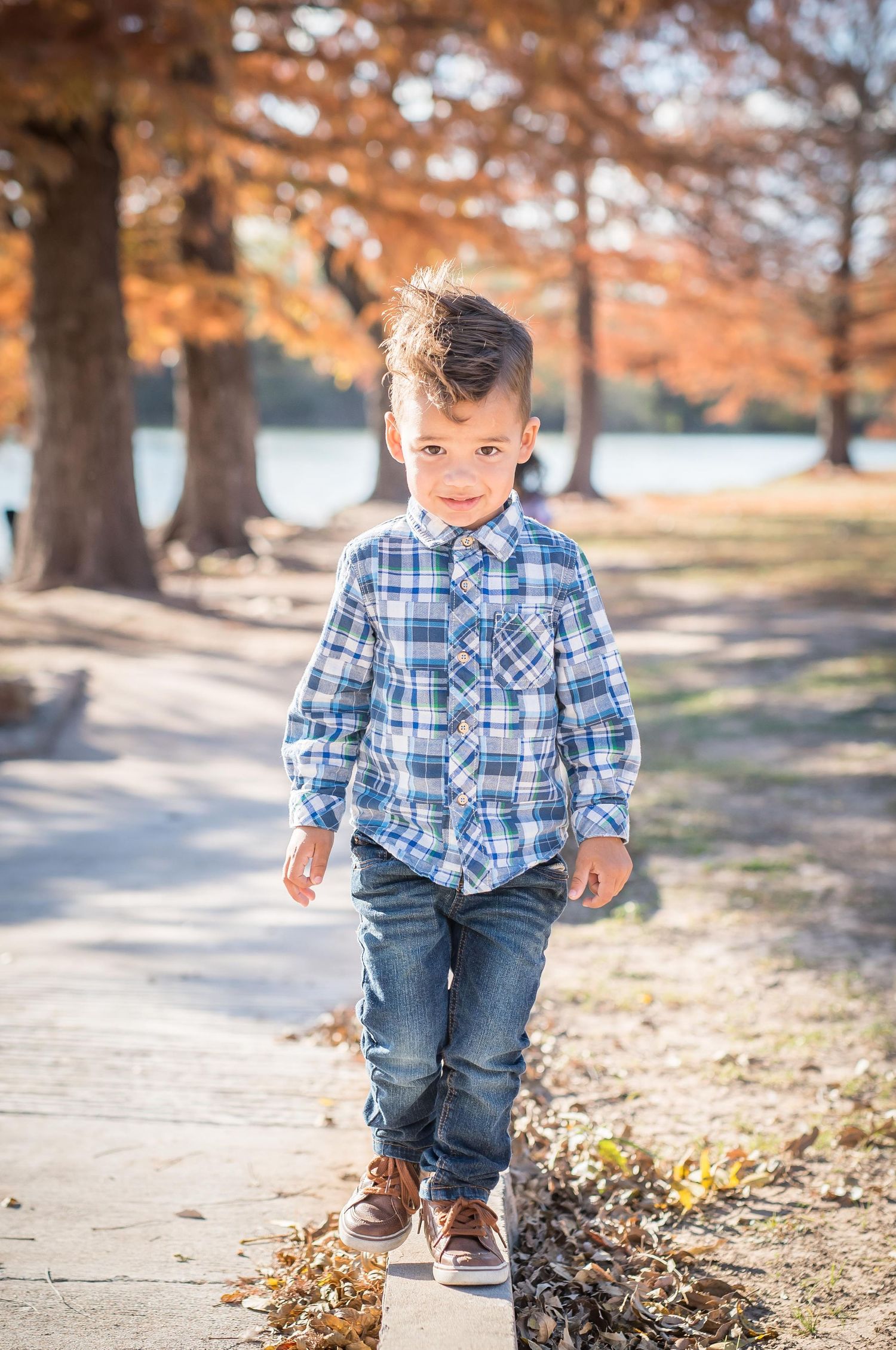 Young boy in plaid shirt walks on path surrounded by autumn trees with orange leaves while taking family photos near Omaha, Nebraska.