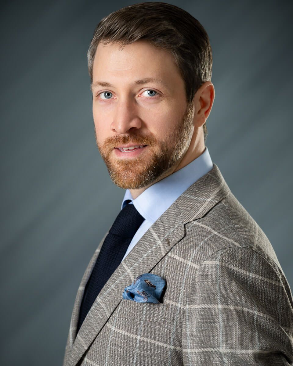 Professional man with beard in plaid suit, blue pocket square, and navy tie poses for formal business headshot.