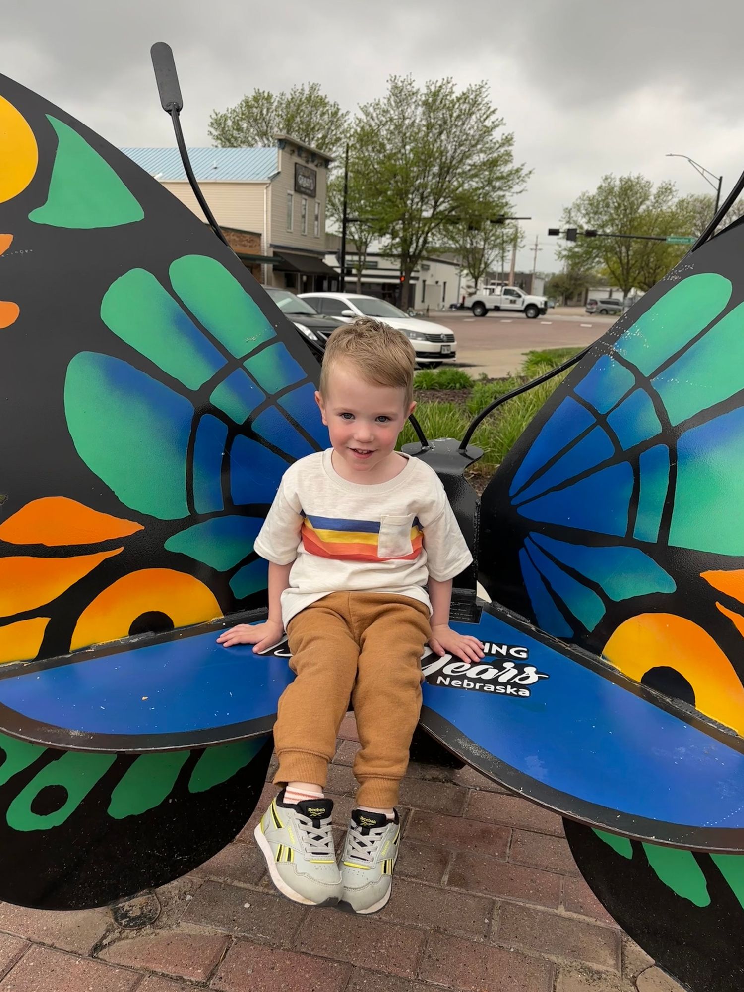 A child sits on a large decorative butterfly bench with vibrant blue and green painted wings outdoors.