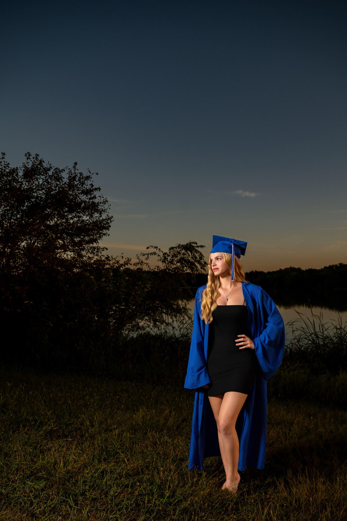 Young woman in blue graduation cap and gown standing outdoors near lake at dusk in dramatic lighting.