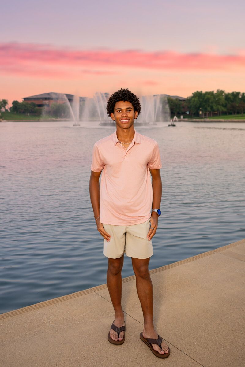 Young man in pink polo and khaki shorts stands by a fountain lake under a vibrant pink sunset sky.