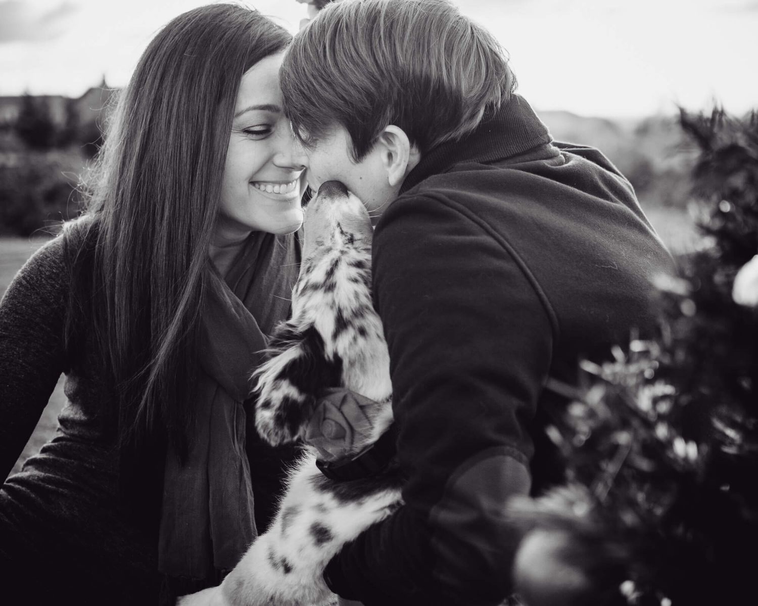 A couple embraces outdoors while a small dog joins in the tender moment in black and white photography.