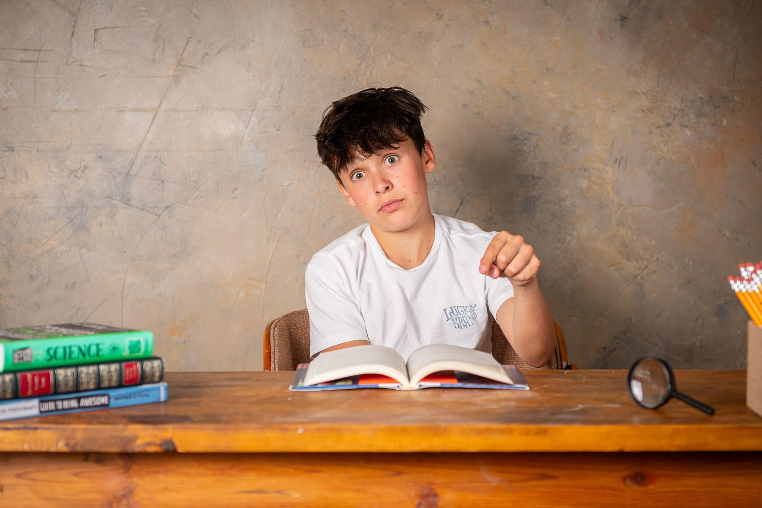 Student in white t-shirt sits at wooden desk with textbooks and clock while studying in room with textured wall.