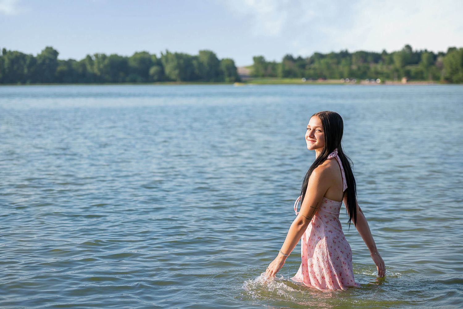 Person in pink dress wading through calm lake waters on a sunny summer day while surrounded by green trees.