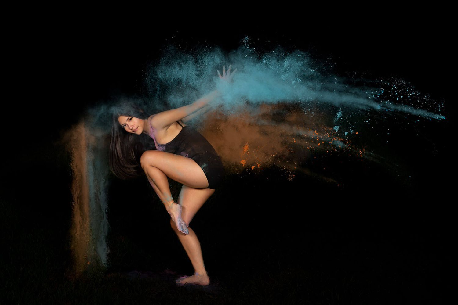 Dancer in black leotard creates dynamic pose with blue and orange powder effects in dark setting.