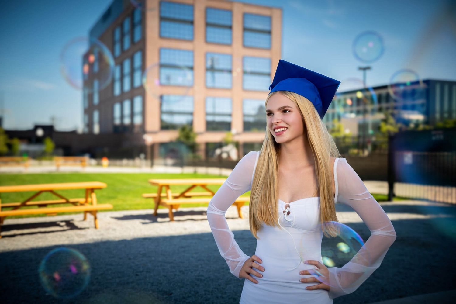 Graduate in blue cap and gown poses outdoors holding diploma against blurred background.
