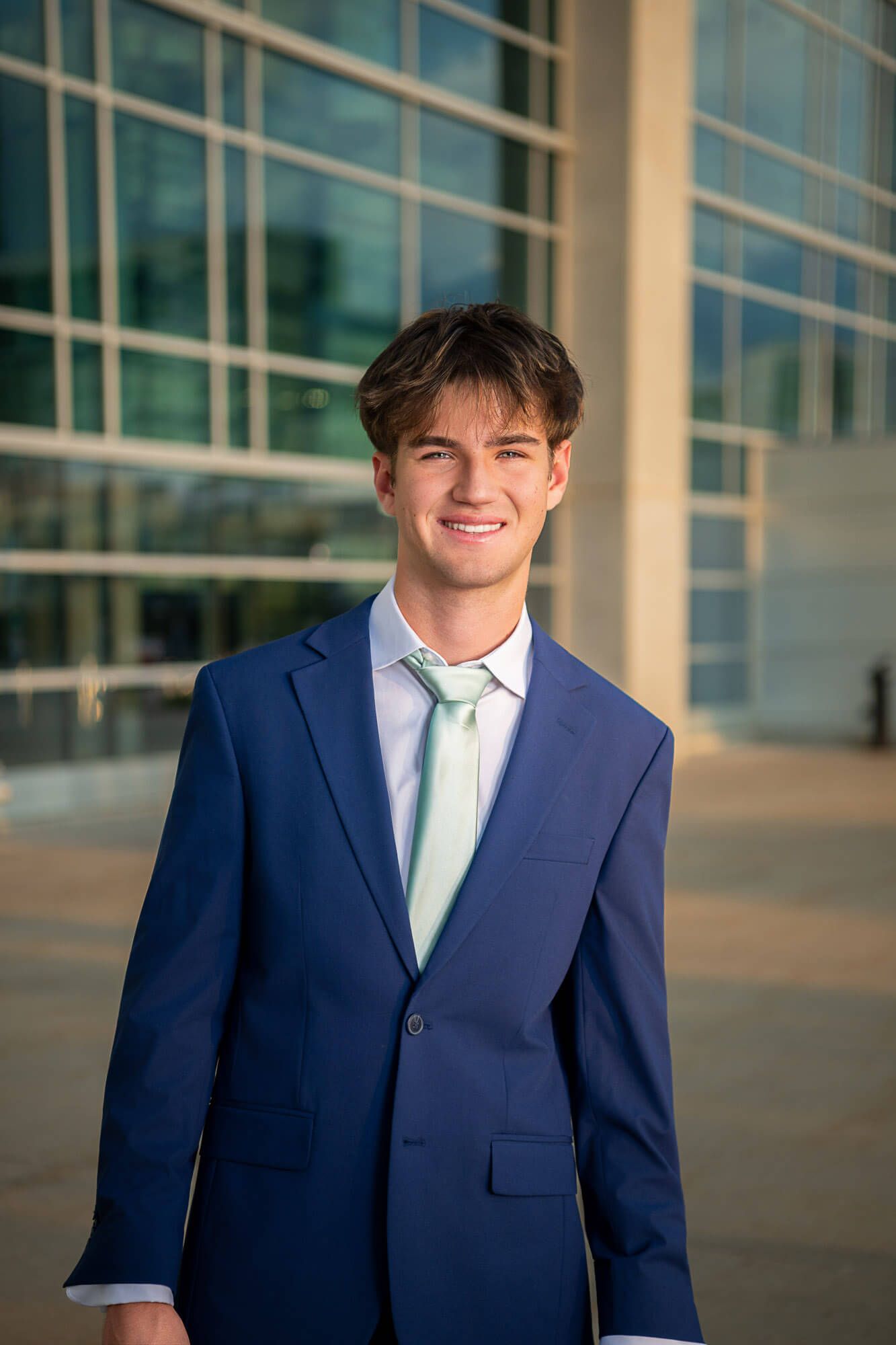 Professional portrait of someone in a navy blue suit and light blue tie standing against a modern building exterior.