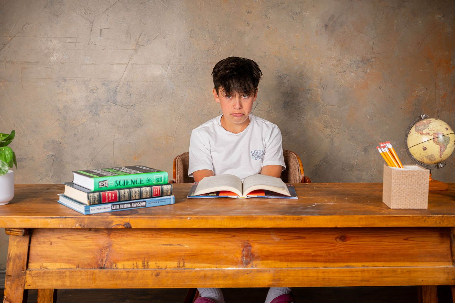 Student sits at wooden desk reading book with textbooks and globe nearby in study setting.
