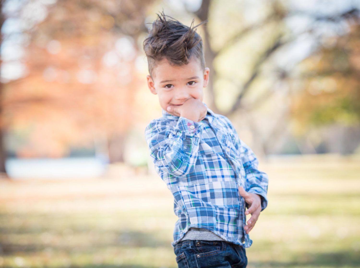 A boy wearing a blue plaid shirt poses playfully in an outdoor setting with autumn trees in the background during family photography session