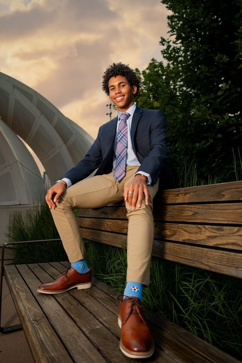 Young man in navy blazer and tan chinos sitting on wooden bench at sunset.