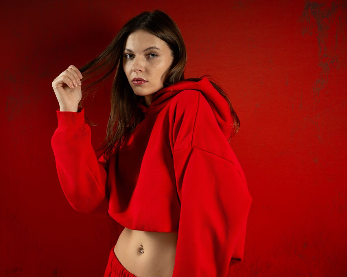 Young woman in red cropped hoodie poses against red background, showing midriff with belly piercing.