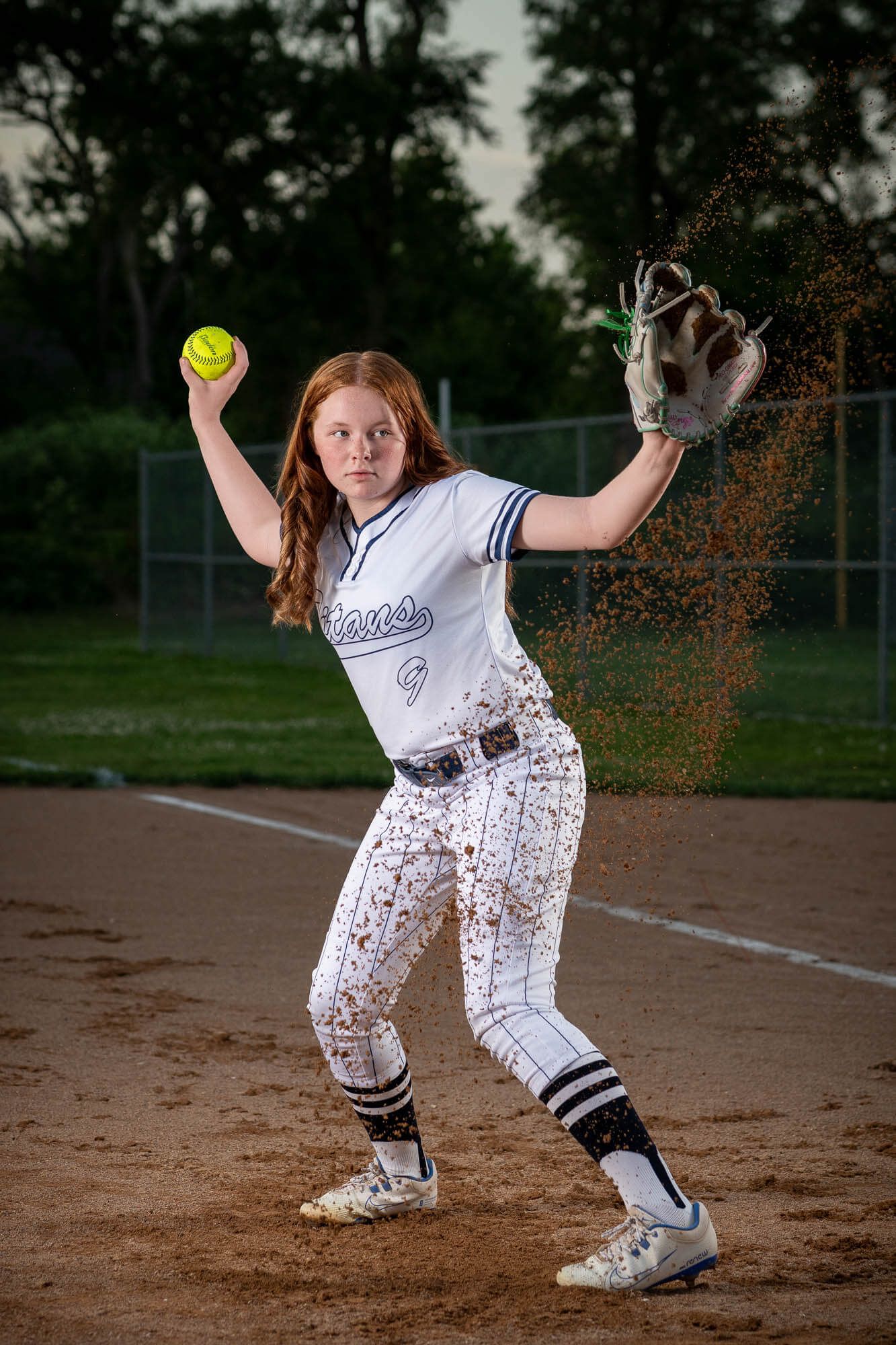 Young female softball player in white uniform winds up to throw ball on dirt field.