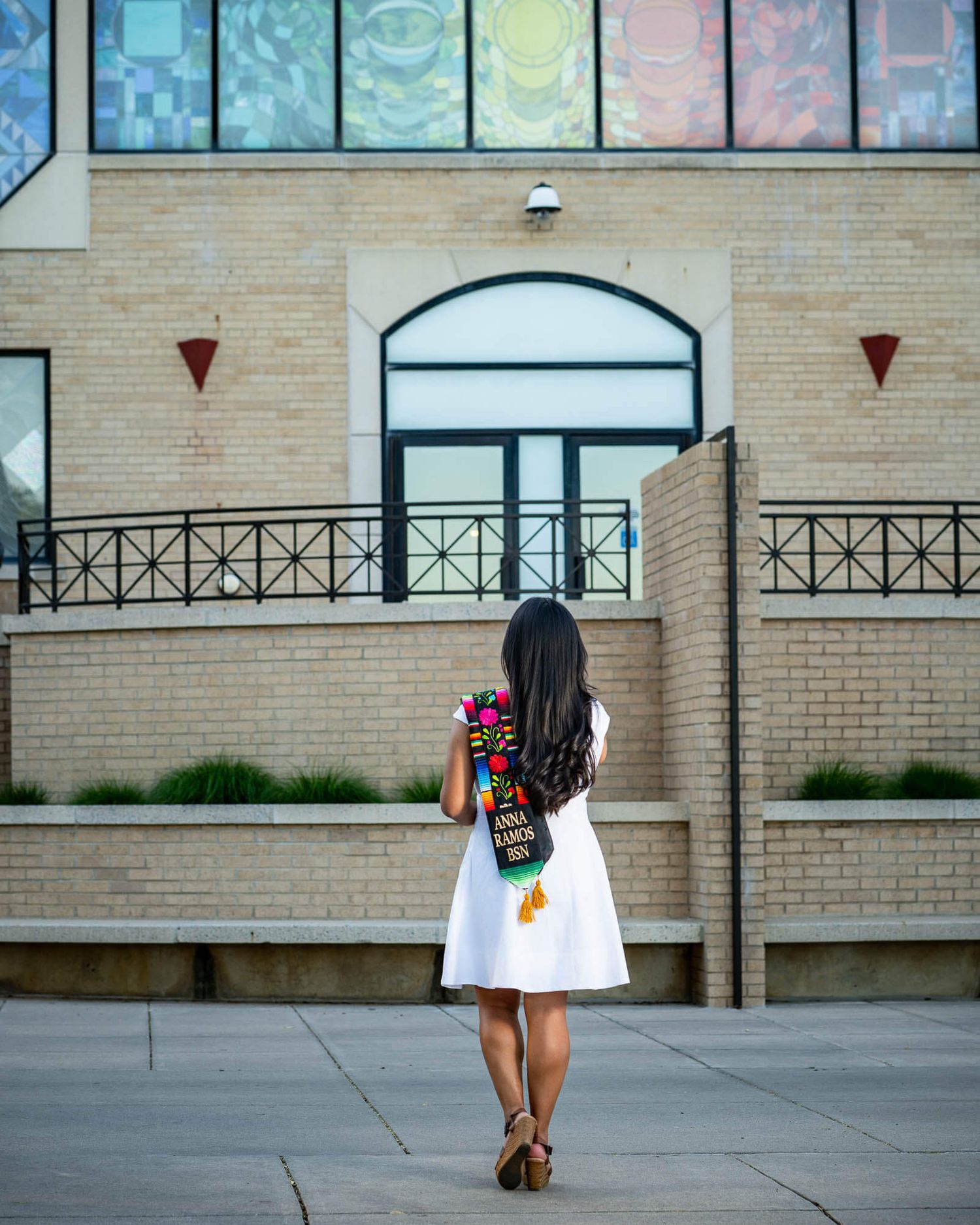 A graduate in a white dress holds a colorful graduation stole while standing before a brick building with stained glass windows.