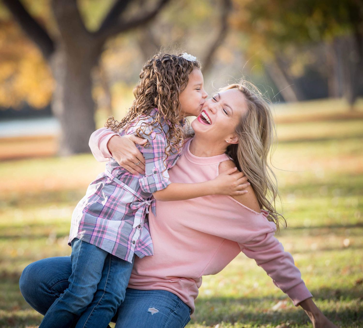 Playful outdoor autumn photoshoot showing joyful embraces and laughter on grassy ground with fall foliage in background.