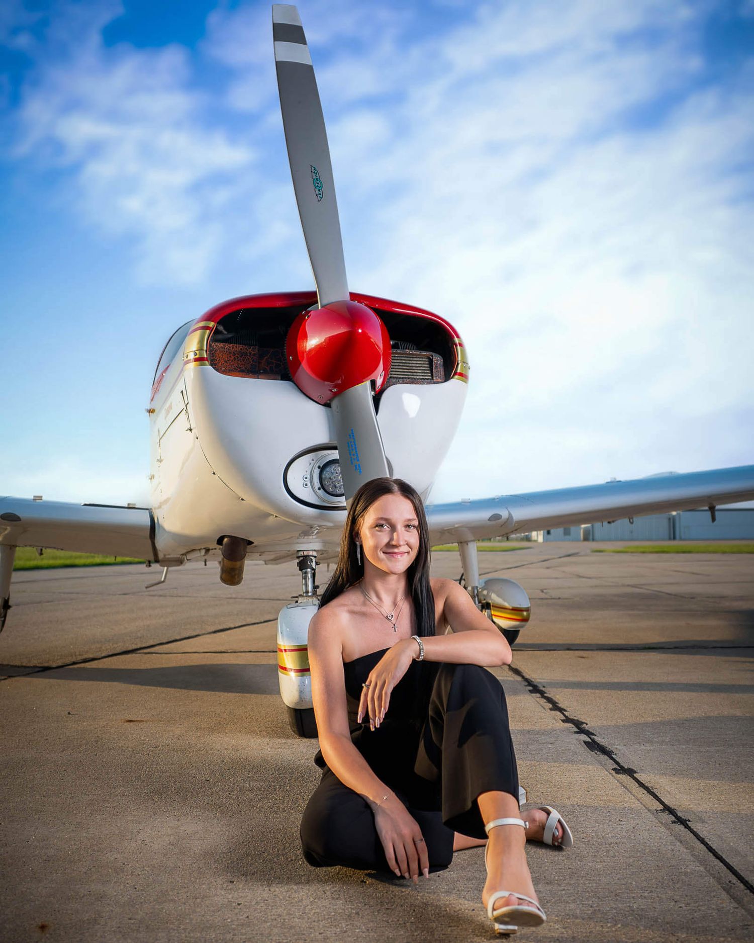 Senior photography near Omaha of a female in a black pantsuit sitting in front of an airplane and blue cloudy sky.
