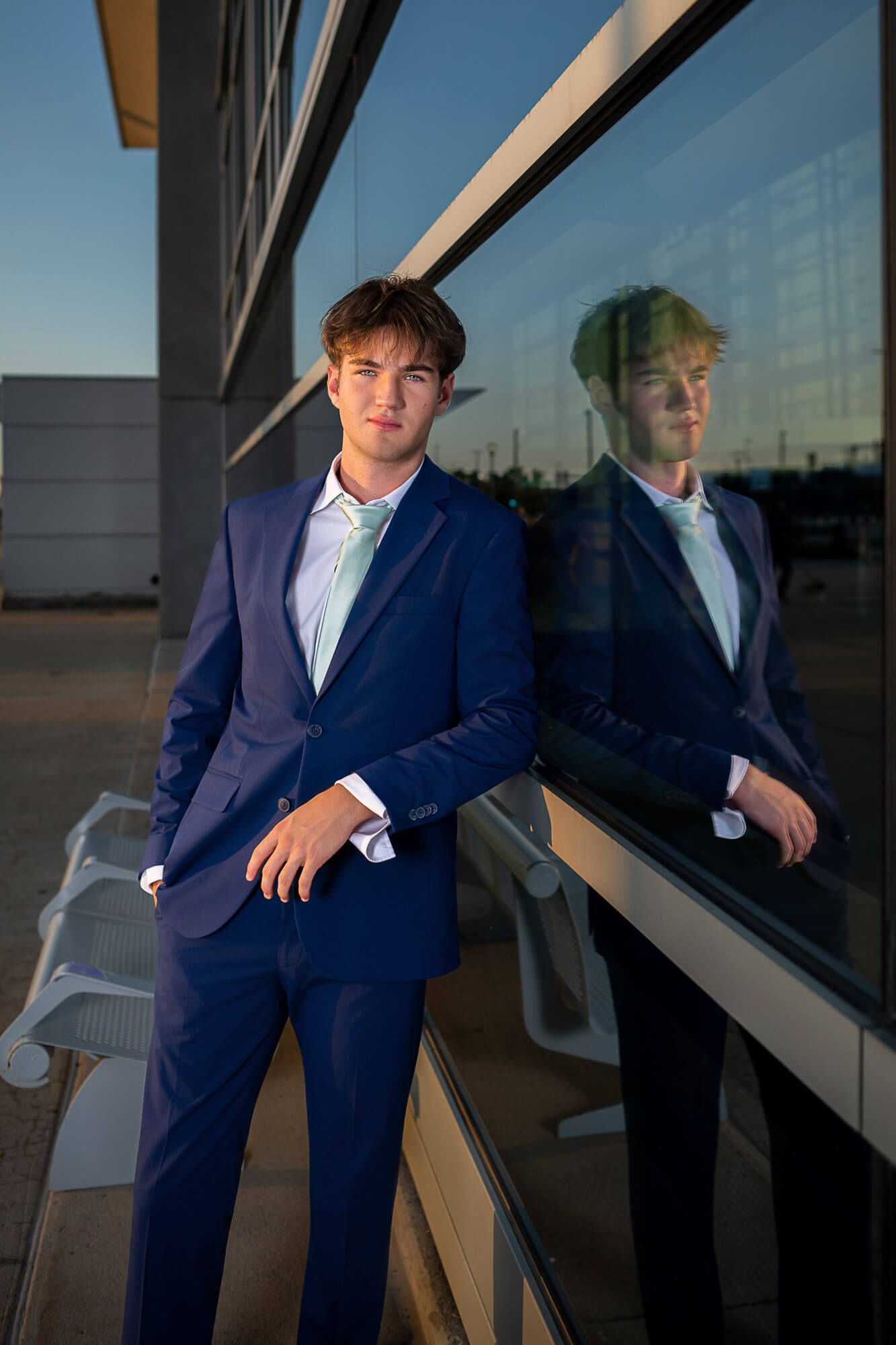 Young man in navy blue suit leaning against glass wall with reflection at sunset.