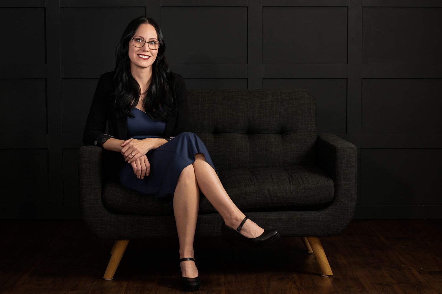 Professional portrait of someone seated on grey couch wearing black blazer in dramatic lighting.