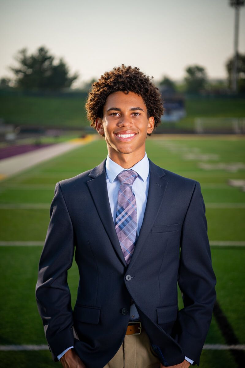 Smiling young man in navy suit and plaid tie posing on football field bleachers.