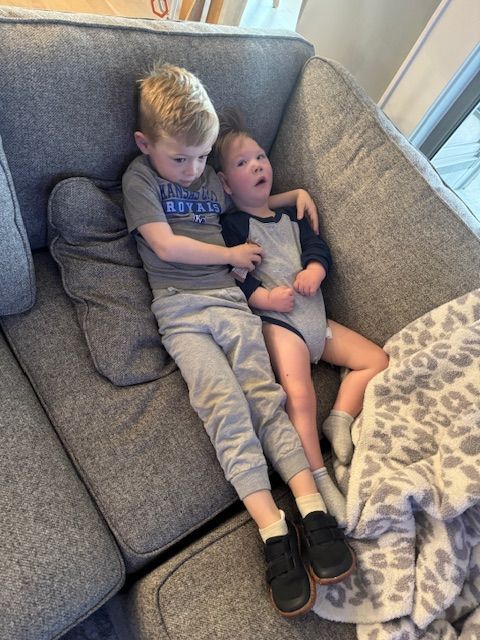 Two children playfully lounging together on a gray couch in a casual, candid moment at home.