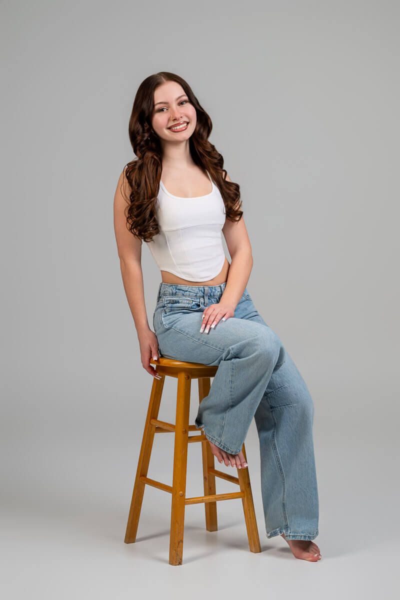 Smiling young woman with curly hair sitting on a wooden stool, wearing a white crop top and wide-leg jeans.