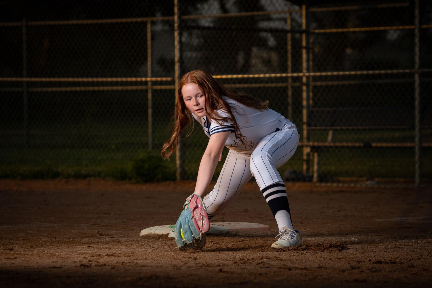 Young female softball player fielding a ground ball at first base during a night game on a dirt infield.