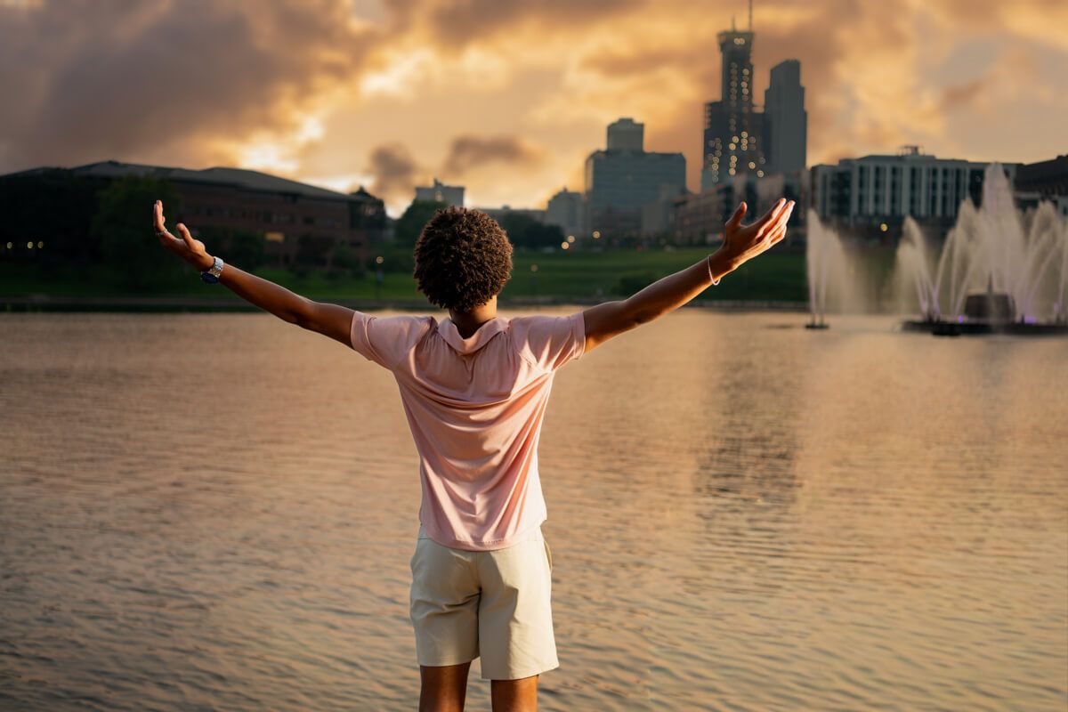 Person standing with arms outstretched facing a city skyline with fountains and a lake at sunset.