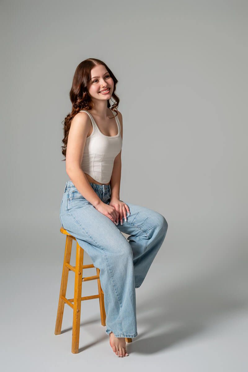 Young woman with curly brown hair sitting on wooden stool, wearing white tank top and light blue jeans, smiling.