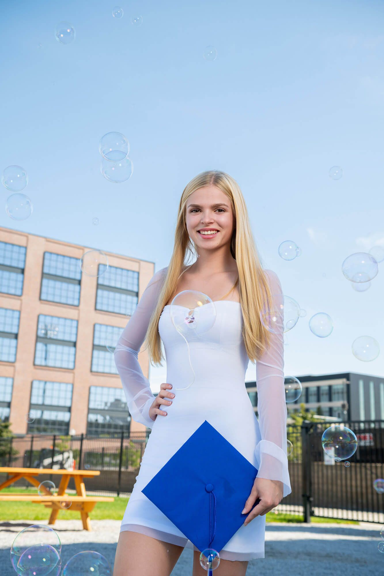 Smiling blonde woman in white dress holding blue graduation cap outdoors with soap bubbles floating around her.