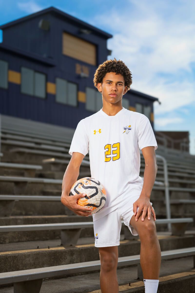 Young soccer player in white Under Armour jersey number 23 holding ball on stadium bleachers.