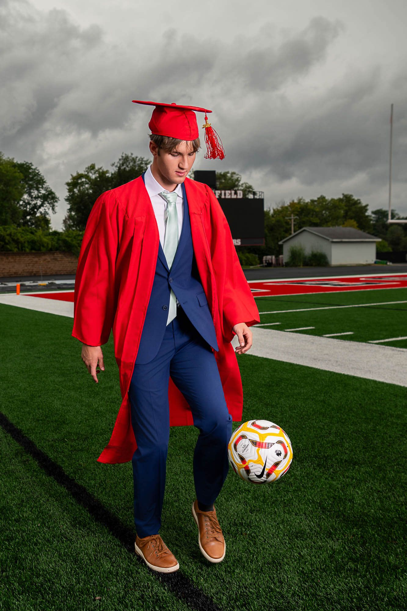 Graduate in red cap and gown with blue suit stands on football field holding soccer ball under stormy sky.